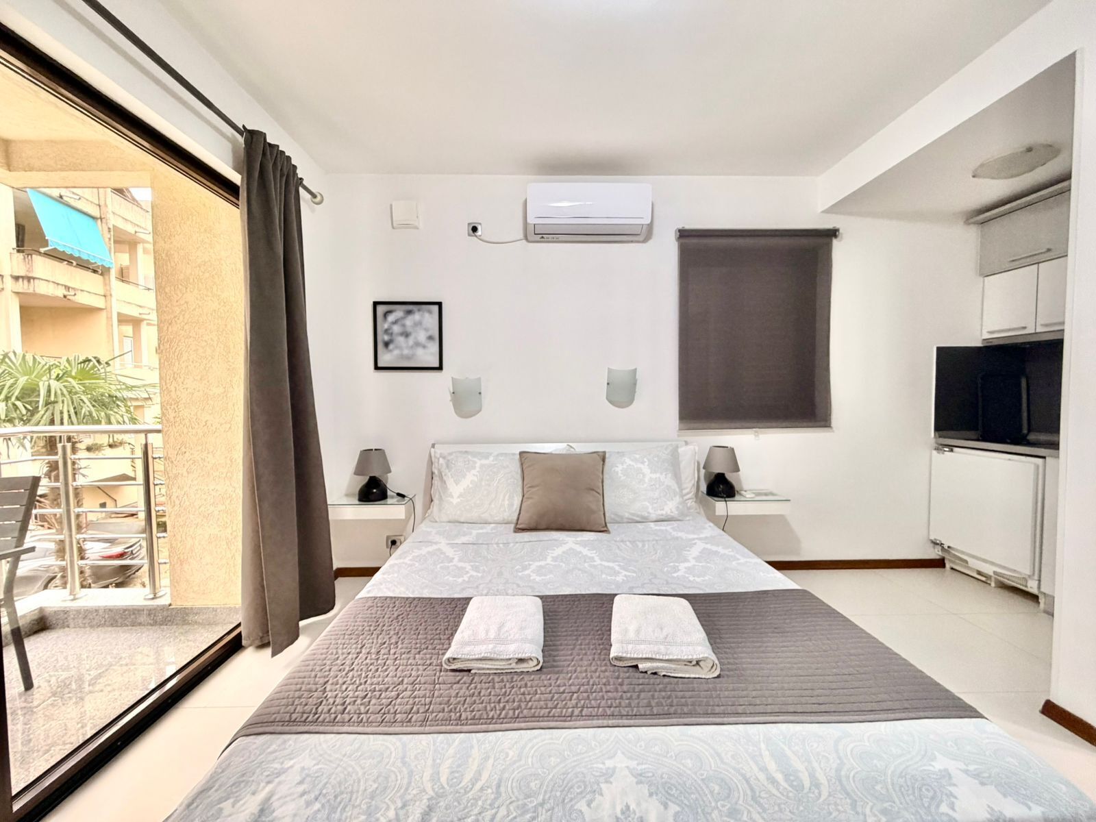 A hotel room with a large bed, a TV, a kitchenette, and a balcony with a view of the outside.