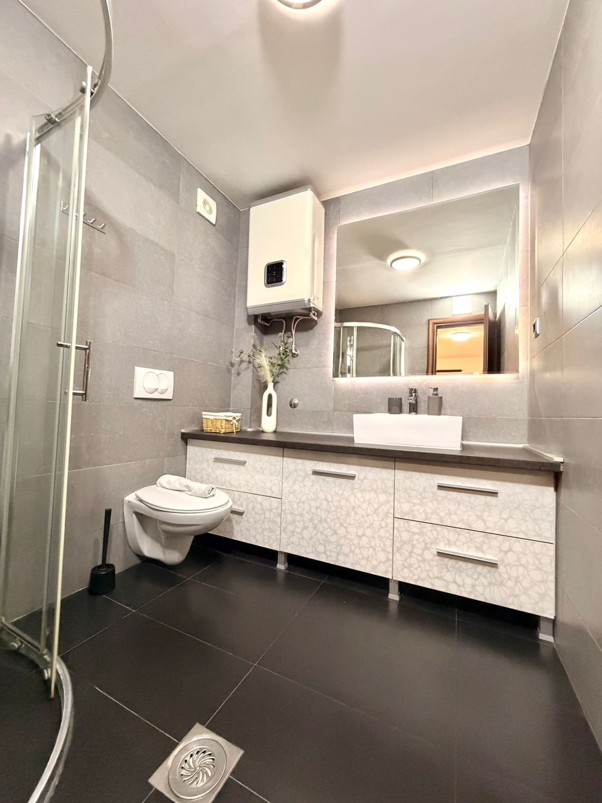 Een moderne badkamer met een douchecabine van glas, een wit toilet, een wastafel met een grote spiegel en een donkere betegelde vloer.
