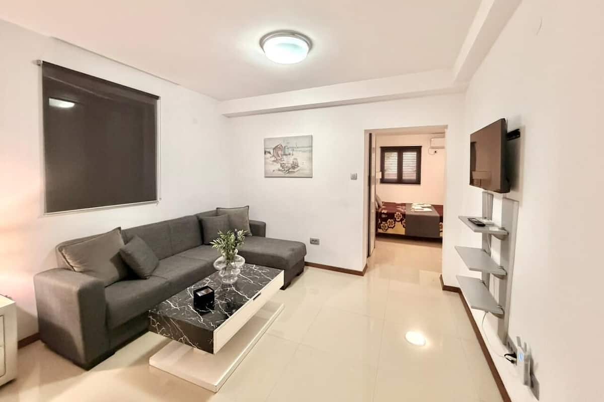 A modern living room with a grey sofa, a coffee table with a vase, a flat-screen TV mounted on the wall, and a hallway leading to a dining area.