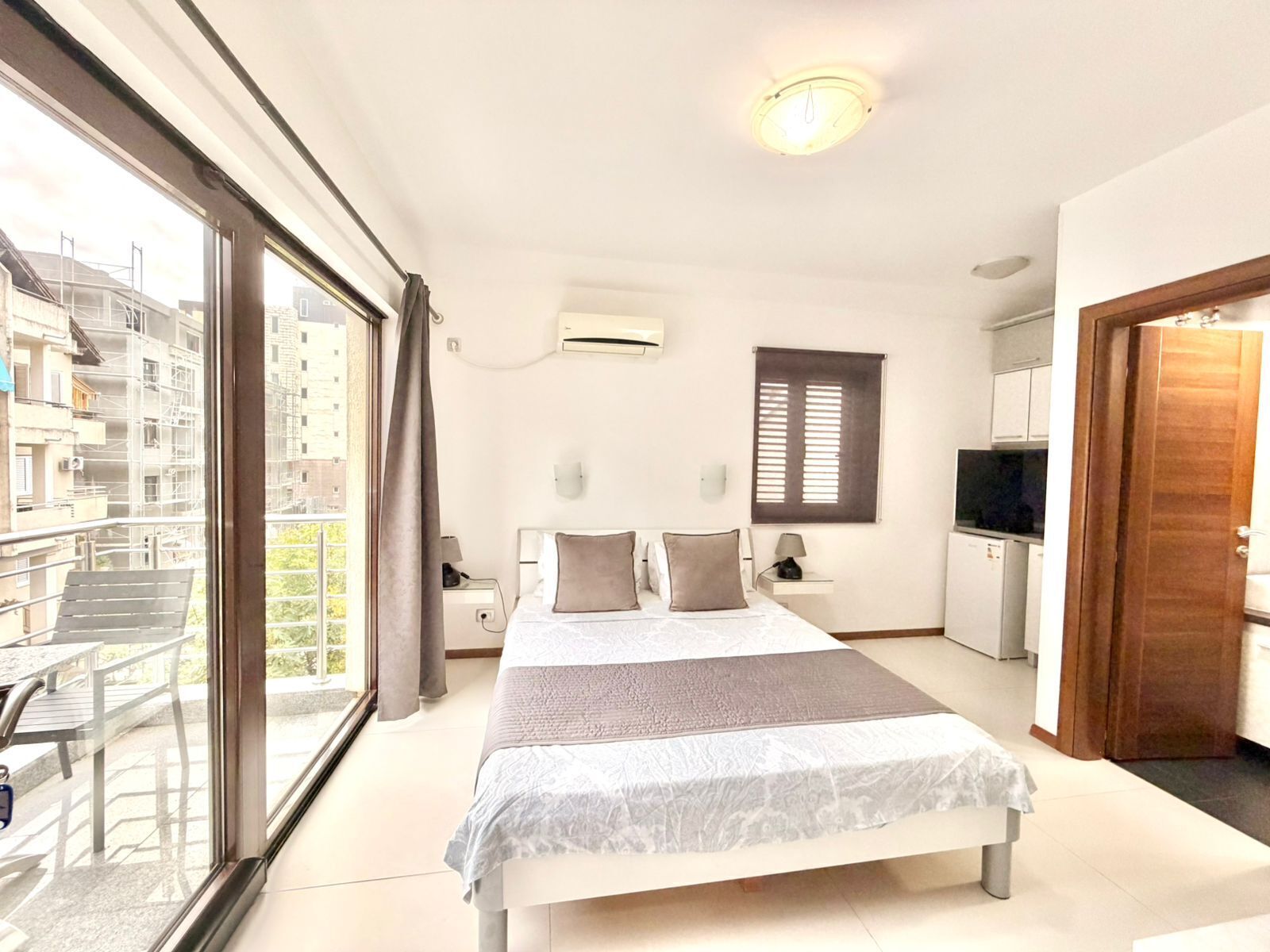 A bedroom with a bed, a TV, a desk, and a window overlooking a balcony.
