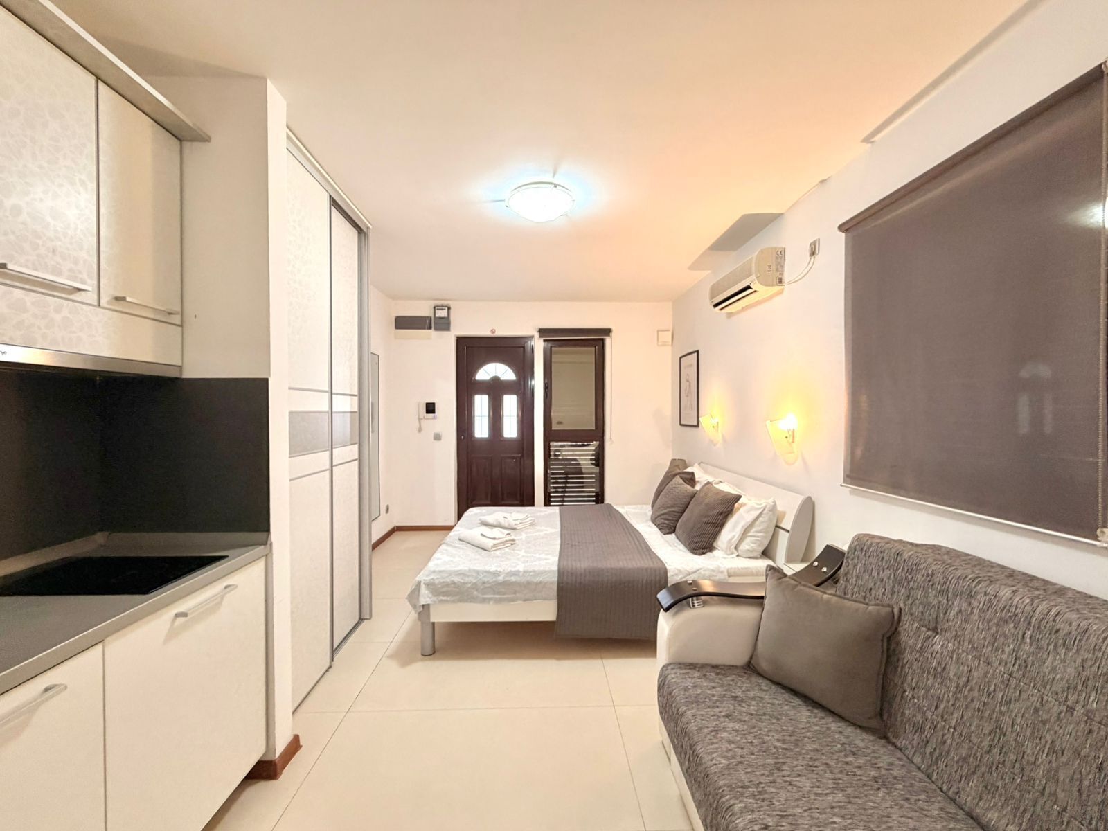 Une chambre d’hôtel compacte avec une kitchenette, un lit doté d’une tête de lit grise, un canapé gris et une grande fenêtre.