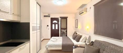 Une chambre d’hôtel compacte avec une kitchenette, un lit doté d’une tête de lit grise, un canapé gris et une grande fenêtre.