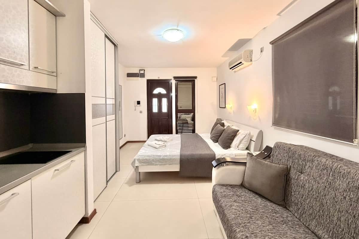 A compact hotel room with a kitchenette, a bed with a gray headboard, a gray sofa, and a large window with a view.
