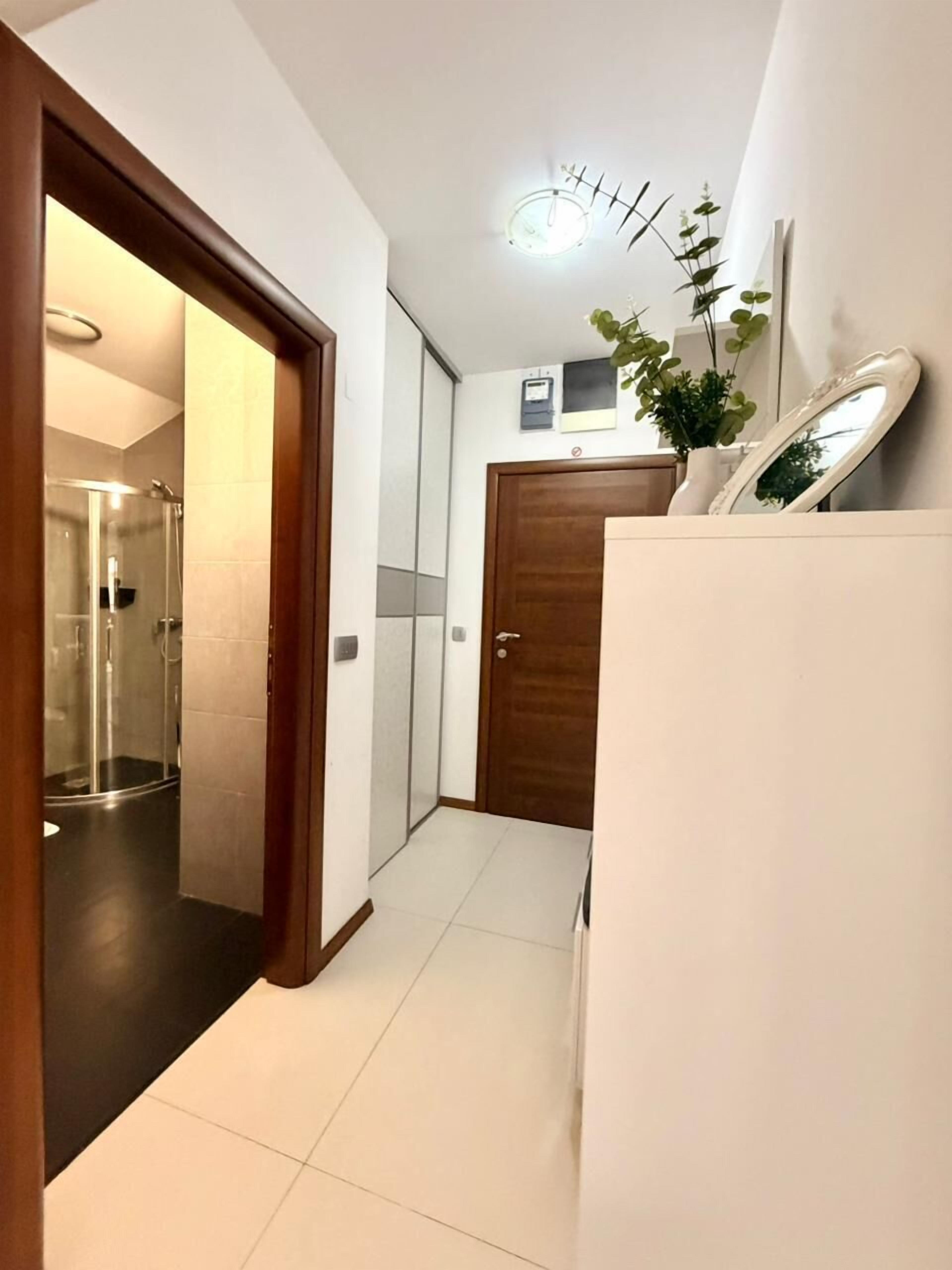 Un couloir avec une porte en bois, un mur blanc, une petite plante en pot et un miroir sur une commode blanche.