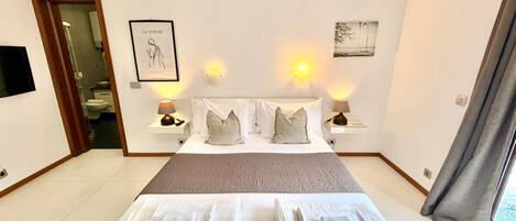 A hotel room with a large bed, two bedside tables with lamps, a framed artwork on the wall, and a view of the outside through a window.