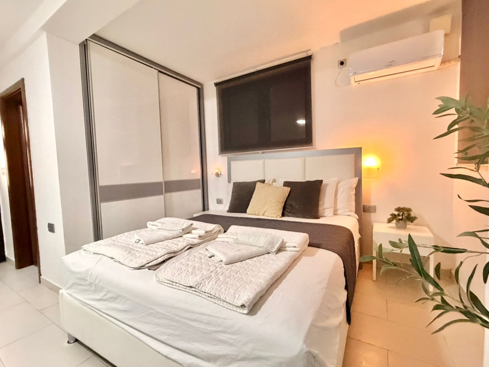 A modern bedroom with a large bed, a sliding door, an air conditioning unit, and a bedside table with a lamp.
