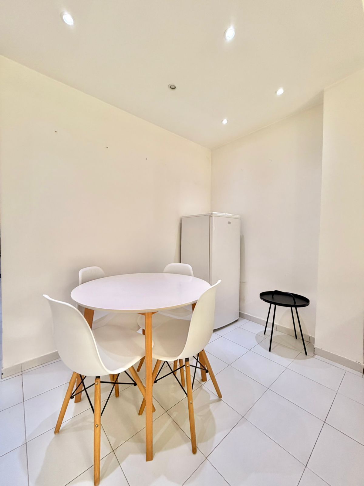 A room with a round table and four chairs, a small black table, and a refrigerator.