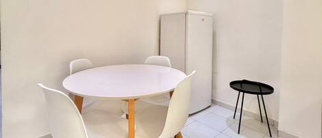 A room with a round table and four chairs, a small black table, and a refrigerator.