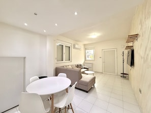A modern living room with a sofa, a dining table with chairs, a TV mounted on the wall, and a door. - Adriatic Urban Apartments (Budva)