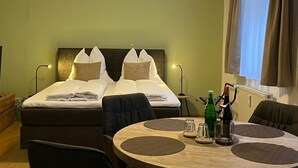 Habitación de hotel con cama, dos mesitas de noche, una mesa redonda con sillas, lámpara colgante y ventana con cortinas.