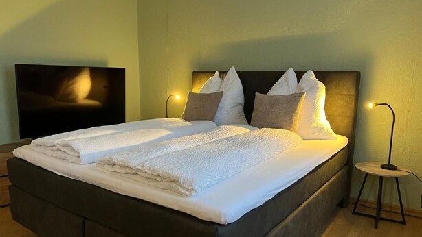 Una cama con ropa de cama blanca y almohadas, cabecera oscura y dos lámparas de noche sobre un piso de madera.