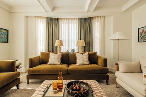 Ein Wohnzimmer mit einem braunen Sofa, zwei Lampen, einem Couchtisch mit einer Schale Feigen und einer Stehlampe.