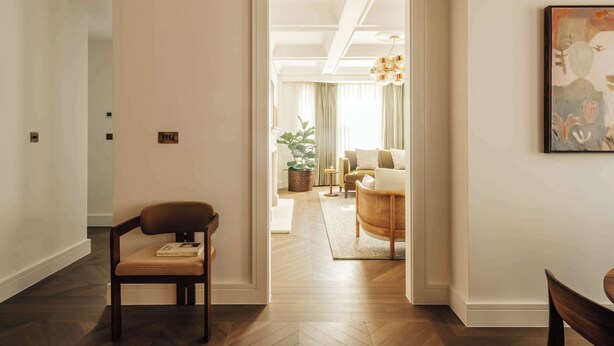 A modern interior with a wooden floor, a brown chair, a white wall, and a doorway leading to a living area with a sofa and a plant. - Mayfair Luxury 3BR at SLO Claridge House (London)