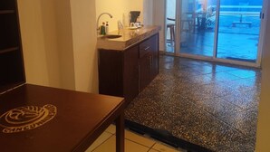 A kitchen with a sink, cabinets, and a countertop. There is a dining table in the foreground. The room has a tiled floor and a view of an outdoor area through a glass door. - Cancún Hotel Boutique & Hostel (Cancún)