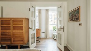 Ein Zimmer mit Holzschrank, einem Stuhl und einem Bett. Eine Tür führt in einen weiteren Raum mit Fenster und Vorhängen.