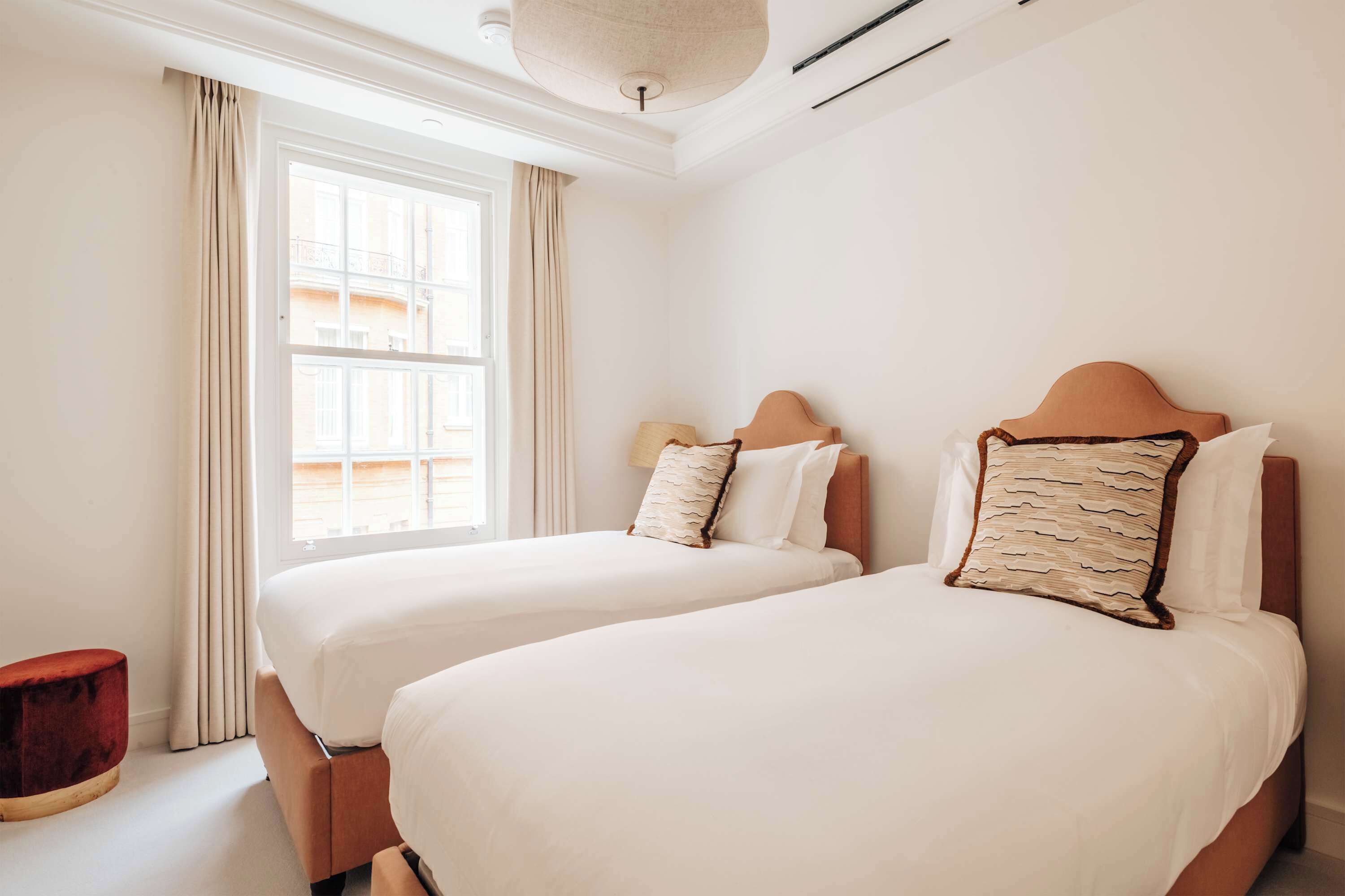 Een hotelkamer met twee bedden, een raam met gordijnen, een rode poef en een plafondlamp.