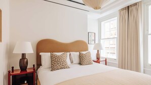 A neatly made bed with a wooden headboard, two bedside tables with lamps, and a window with curtains. - SLO Claridge House: 2BR apartment Mayfair (London)
