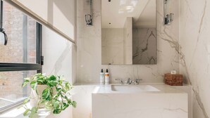 A modern bathroom with a large mirror, white marble walls, a wooden cabinet, and a potted plant by the window. - SLO Claridge House: 2BR apartment Mayfair (London)
