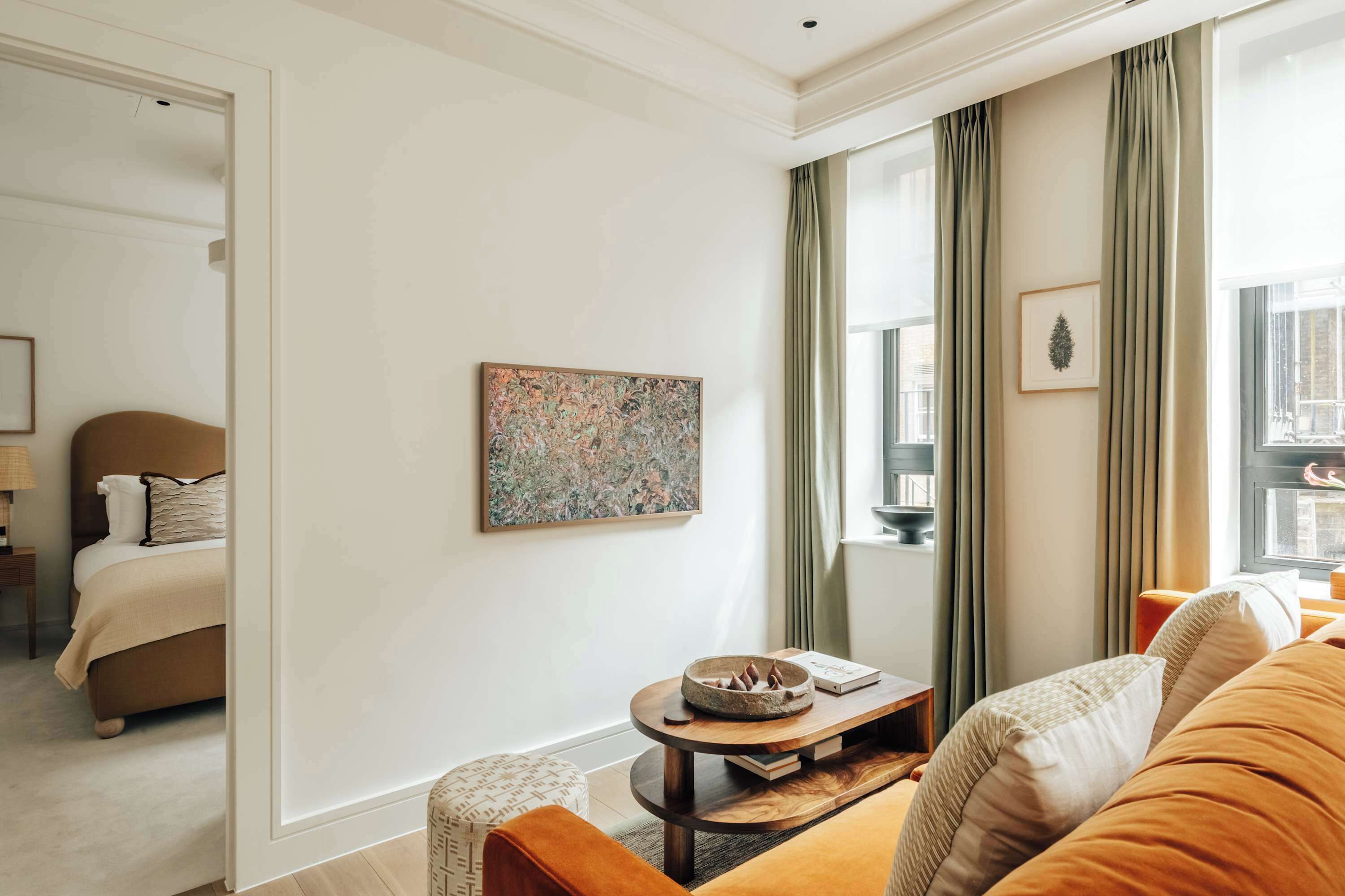 A modern hotel room with a bed, a seating area with a coffee table, a painting on the wall, and large windows with curtains.