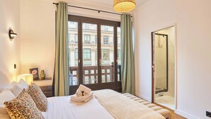 Habitación de hotel con una cama grande, una mesita de noche con lámpara, vista a un edificio a través de una puerta de cristal y un balcón con cortinas verdes.