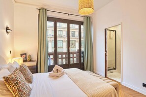 Ein Hotelzimmer mit einem großen Bett, einem Nachttisch mit Lampe, Blick auf ein Gebäude durch eine Glastür sowie ein Balkon mit grünen Vorhängen.