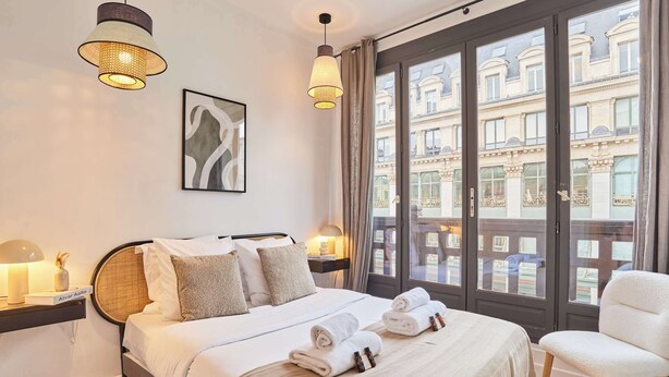 Habitación de hotel con una cama, lámparas de noche, una silla y vistas a edificios a través de ventanas amplias.