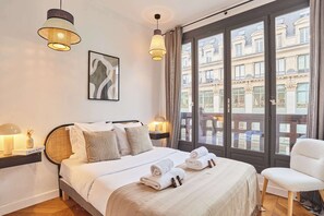 Ein Hotelzimmer mit einem Bett, Nachttischlampen, einem Sessel und Blick auf Gebäude durch große Fenster.