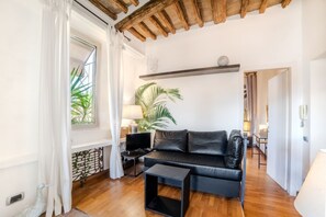 Una sala de estar con techo de madera, un sofá de cuero negro, una mesita pequeña negra, un televisor y una planta grande.