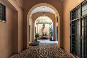 Un corridoio con pavimento in pietra, archi, piante in vaso e una porta con cancello in metallo.
