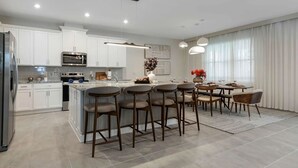 Eine moderne Küche mit weißen Schränken, einer zentralen Kochinsel, Barhocker und einem Essbereich mit Tisch und Stühlen.