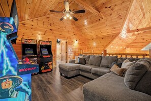 Un interior acogedor de cabaña de madera con un sofá seccional, juegos de arcade y un ventilador de techo.