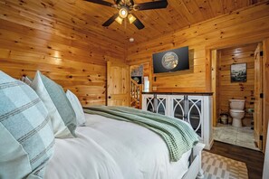 A wooden cabin bedroom with a bed, a ceiling fan, a mounted television, and a bathroom with a toilet and sink. - Firefly Haven Private Indoor Pool, Hot Tub & Game Room Near Dollywood (Pigeon Forge)