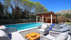 Zona junto a la piscina con sillones de descanso, una mesa de madera con fruta y una estructura de pérgola.