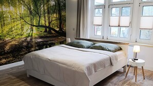 Quarto com cama, mesa de cabeceira, luminária e uma janela grande com persianas.