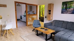 Uma sala de estar moderna com um sofá cinzento, uma mesa de centro de madeira e uma poltrona azul. Uma cozinha fica visível através de uma porta aberta, apresentando uma parede de pedra e armários de madeira.
