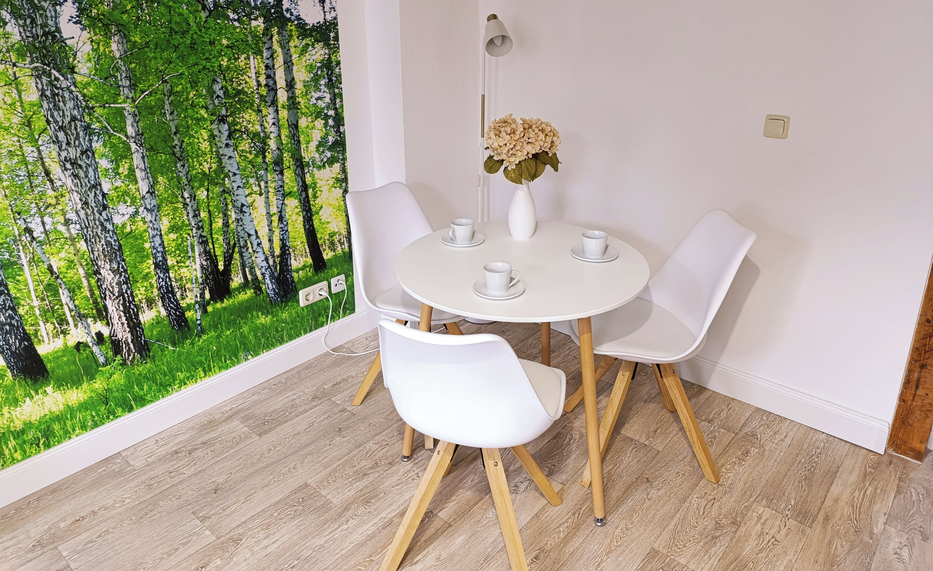 Un coin repas avec une table ronde, quatre chaises blanches, un vase de fleurs et une fresque murale représentant une forêt.