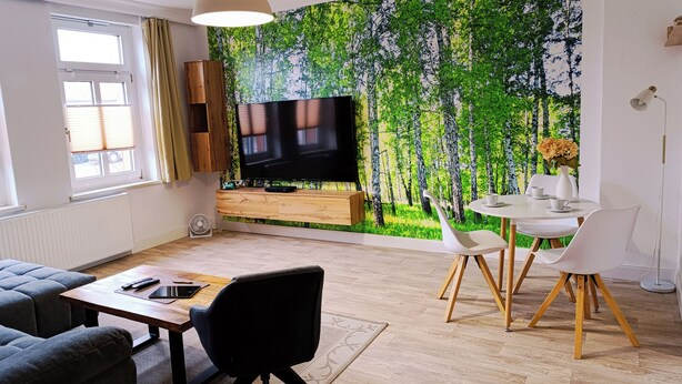 Uma sala de estar moderna com uma televisão de tela plana grande, uma área de jantar com cadeiras brancas e uma mesa de centro de madeira.