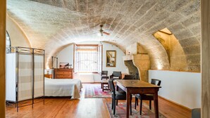 A room with a wooden floor, a dining table with chairs, a bed with a white bedspread, a radiator, and a window with bars. - Guest Room “Room 1” with Shared Pool, Communal Terrace, and Wi-Fi (Vers Pont du Gard)