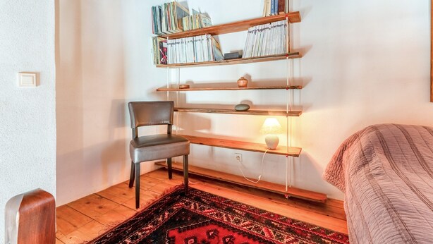 Ein Zimmer mit Holzregal, einem Sessel, einem Bett und einem gemusterten Teppich.