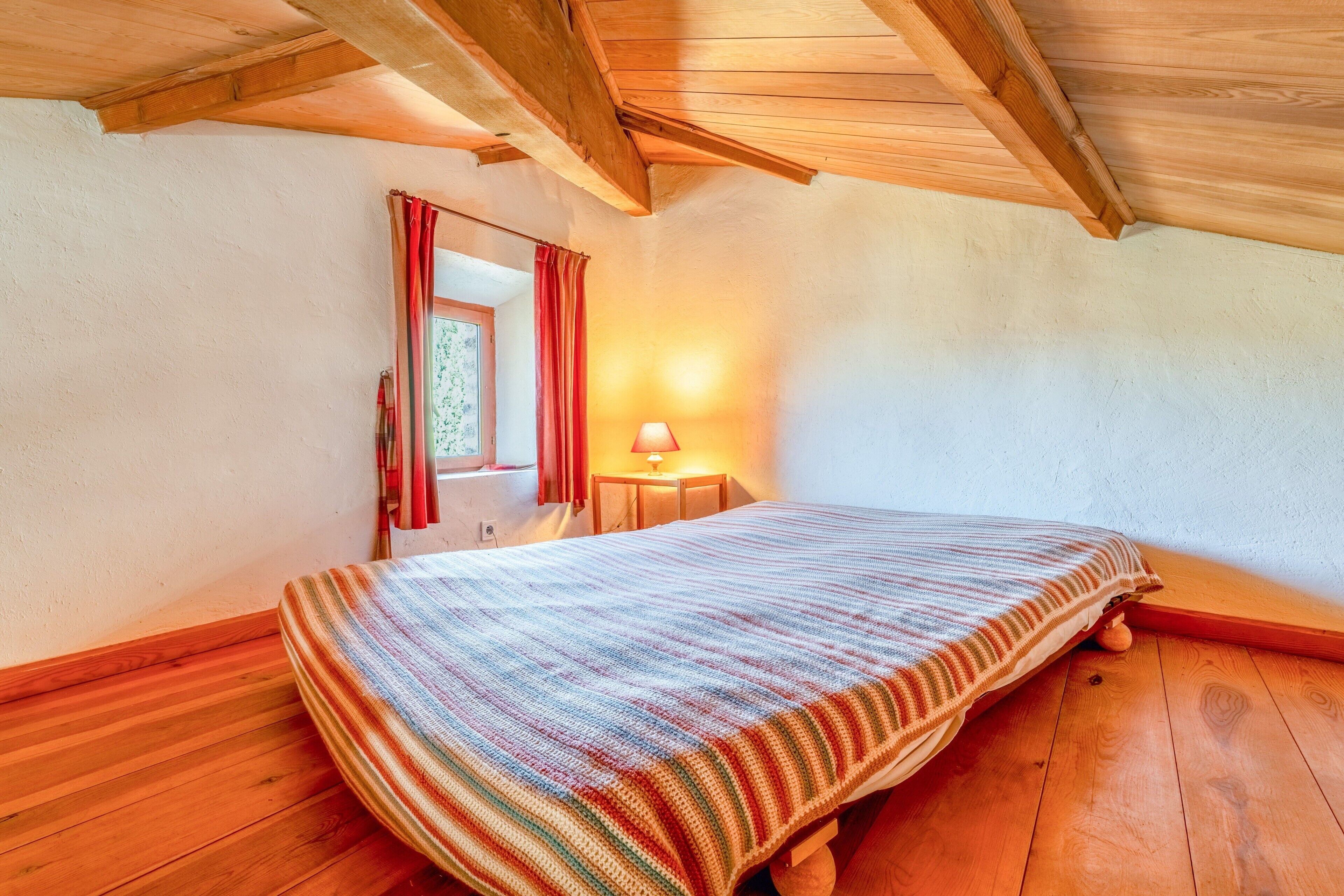 Een kamer met houten vloer, een bed met gestreept dekbed, een raam met rode gordijnen en een tafel met lamp.