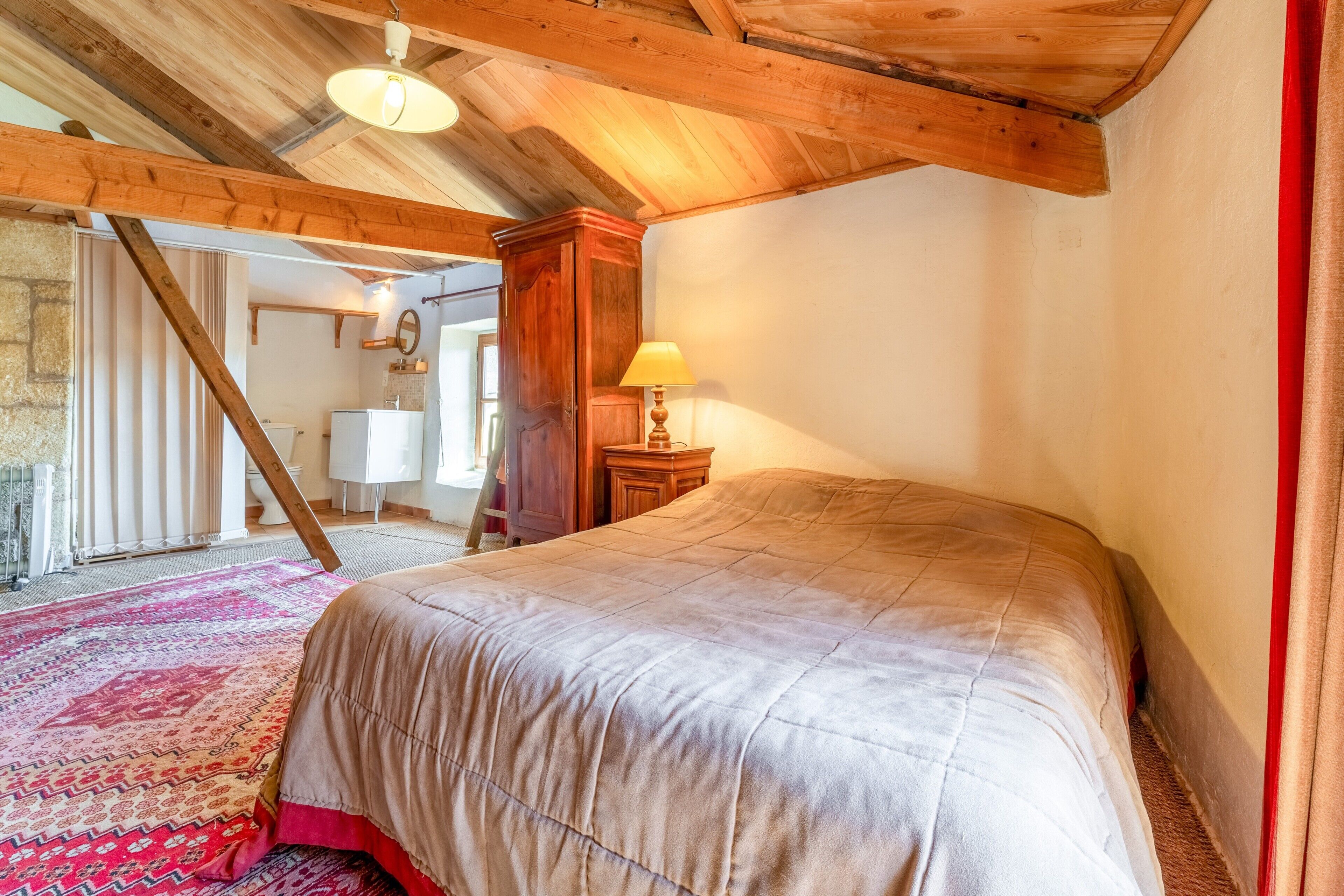 Ein Schlafzimmer mit einem großen Bett, Holzbalkendecke, einem kleinen Nachttisch mit Lampe und einem roten Vorhang.