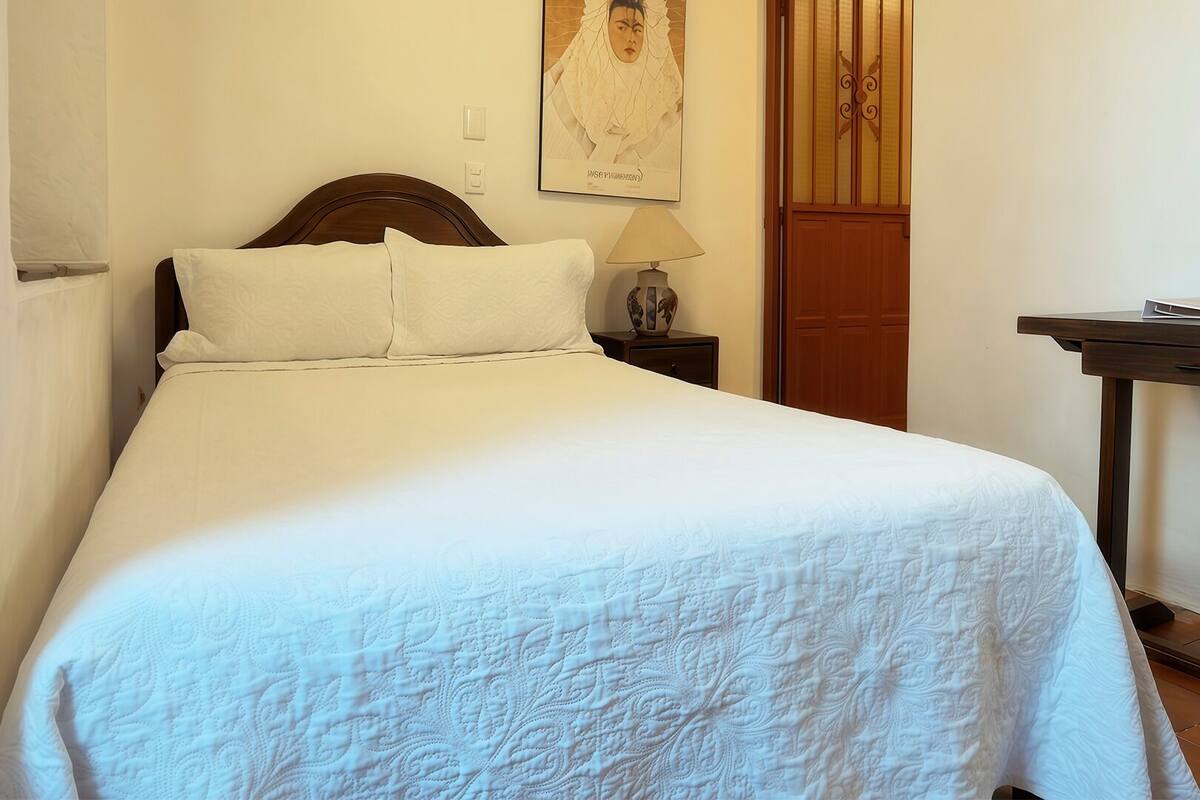 Uma cama bem arrumada com colcha branca, cabeceira de madeira, mesa de cabeceira com abajur e porta de madeira.