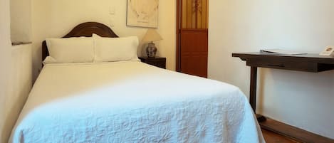 A neatly made bed with a white bedspread, a wooden headboard, a bedside table with a lamp, and a wooden door.