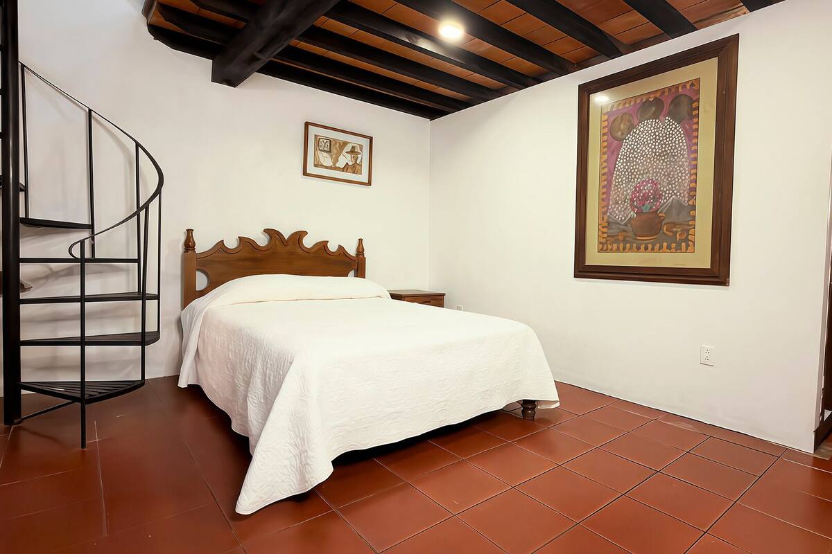 Quarto com cama, escada em espiral, uma grande obra de arte emoldurada e porta de madeira.