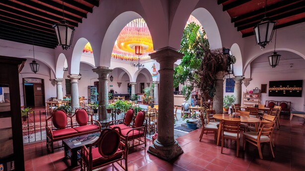 A spacious indoor area with multiple seating arrangements, including red cushioned sofas and wooden tables, surrounded by arches and columns. - Hotel Posada Casa de la Tía (Oaxaca)