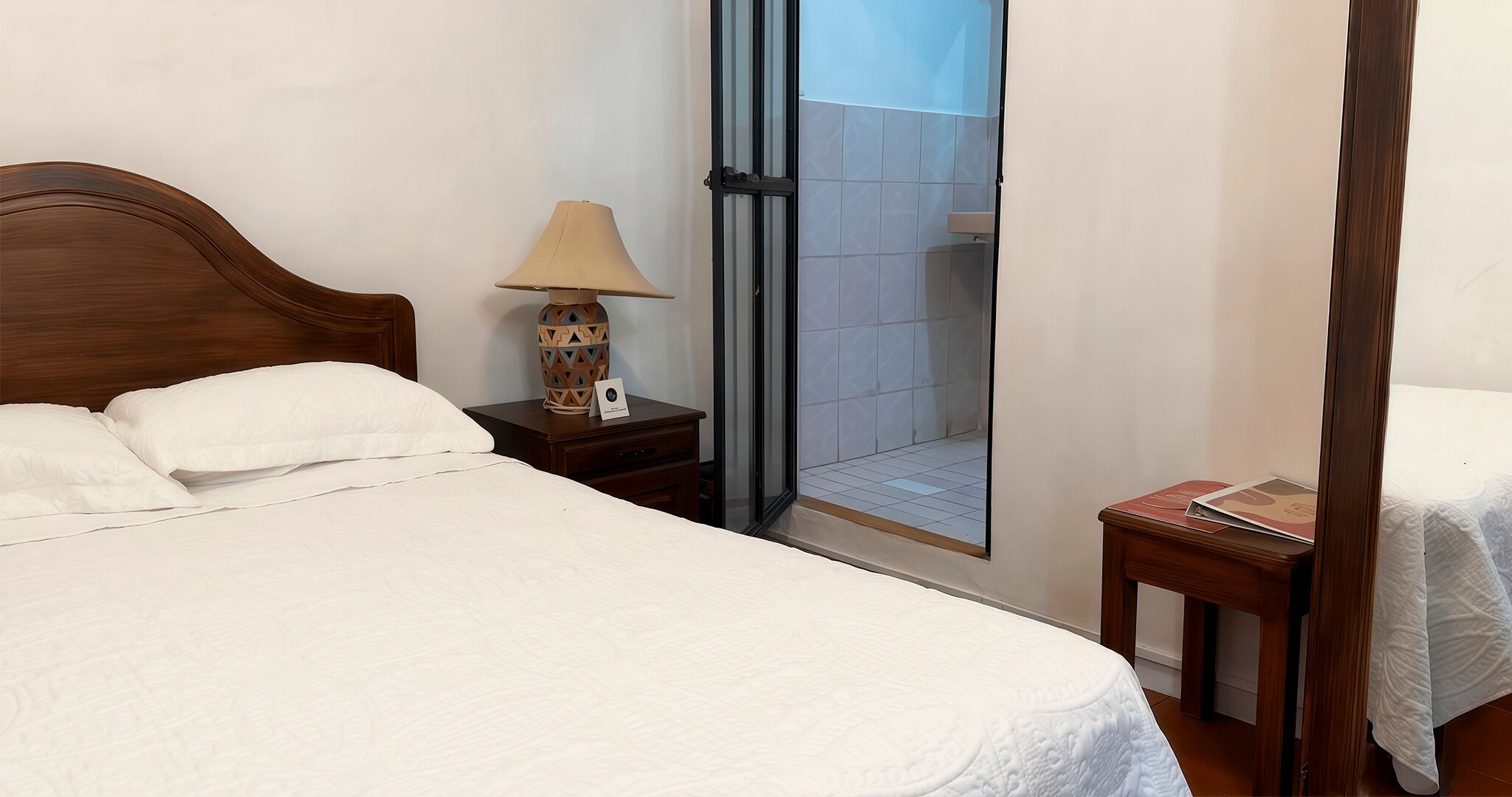 Une chambre d’hôtel avec un lit, une table de chevet avec une lampe, une petite table avec un livre et une porte-fenêtre coulissante donnant sur un espace douche.