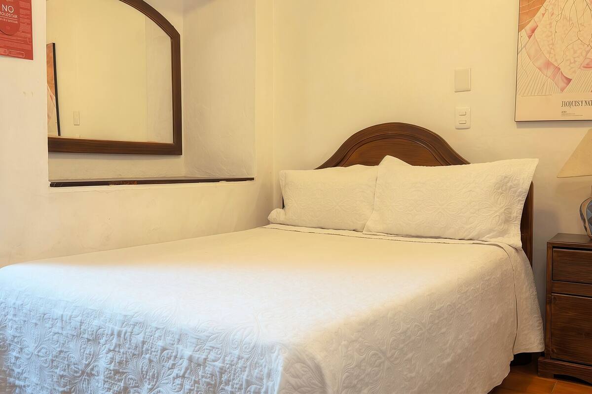 Uma cama bem arrumada com lençóis brancos, cabeceira de madeira, mesa de cabeceira com abajur e um espelho fixado na parede.