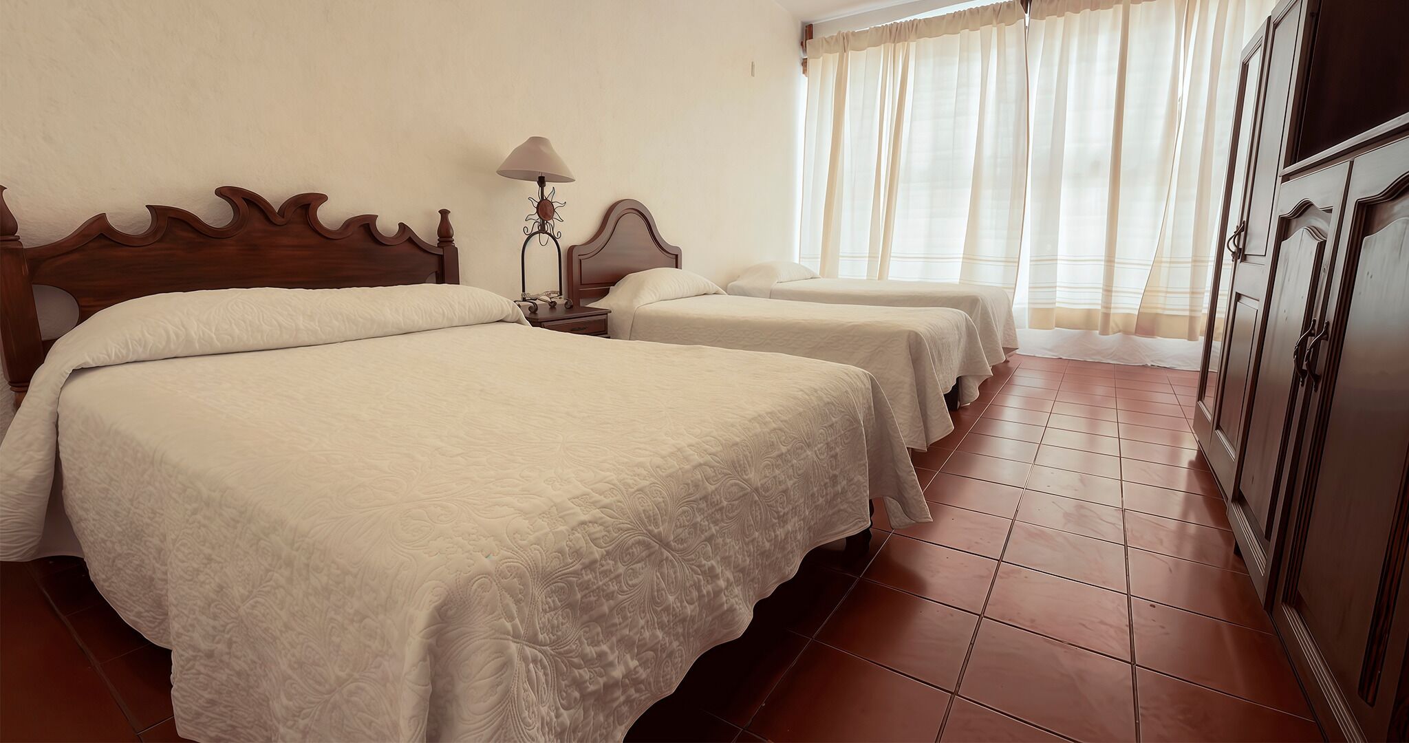 Quarto de hotel com duas camas, uma mesa de cabeceira, uma luminária e um guarda-roupa.