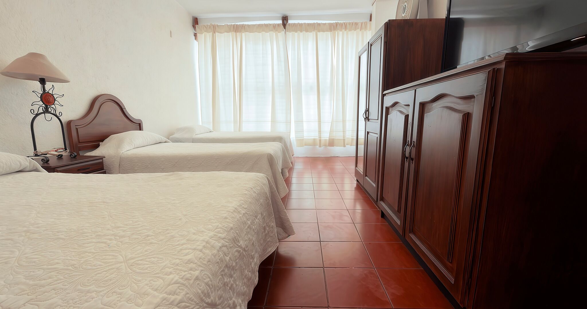Una camera d'albergo con due letti, un armadio in legno, un comodino con lampada e una finestra con tende leggere.