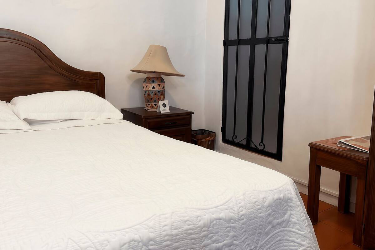 Uma cama bem arrumada com cabeceira de madeira, abajur e janela com ripas pretas.